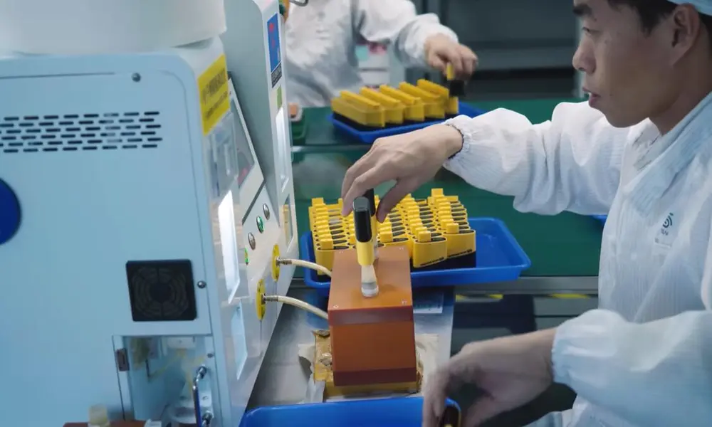 Vape manufacturing quality control: A factory worker in cleanroom attire places a yellow and black electronic cigarette component onto a testing machine on the assembly line, ensuring product standards during production.
