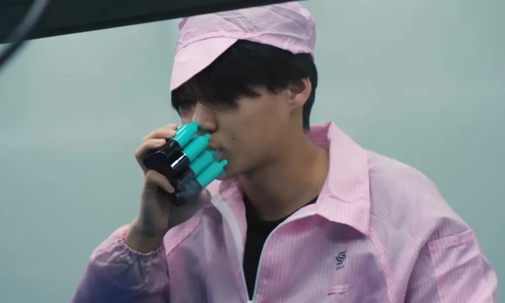 Vape factory quality assurance: A worker wearing a pink cleanroom jacket and cap performs quality control, simultaneously testing a cluster of black and teal electronic cigarette devices by inhaling to check functionality on the production line.